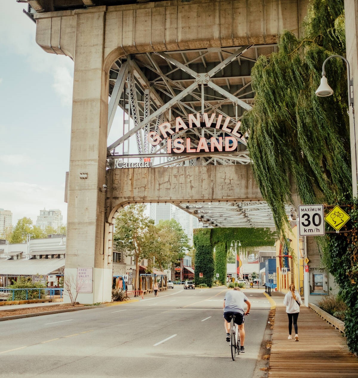 A person riding a bike and person walking on a path underneath a bridge with a sign that says Granville Island. 