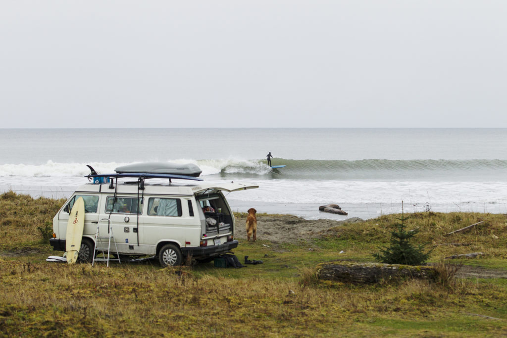 A VW van is parked on the beach, overlooking a surfer riding the waves.