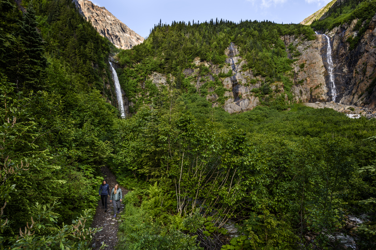 Two waterfalls pour down a rocky mountain and disappear into the trees below. Two women walk along a trail in the foreground.