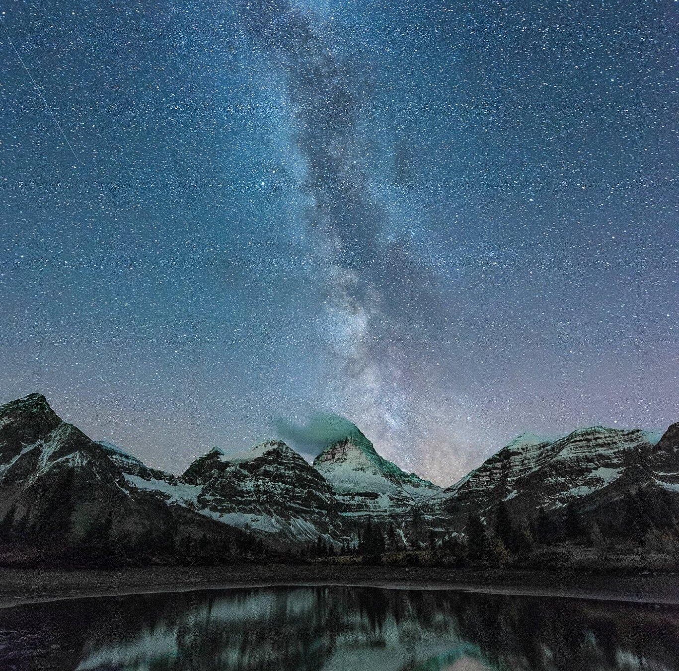 A clear alpine lake is in the foreground with snowy mountain peaks behind. Straight up from the peaks in the centre of the frame is the Milky Way in a star-filled sky.