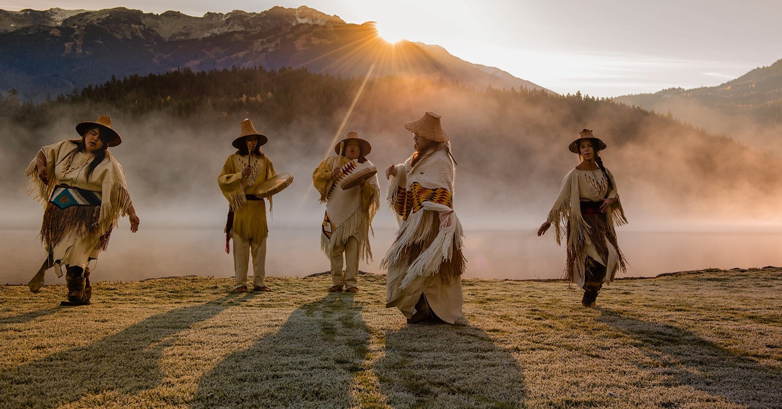 Five Indigenous people in traditional attire dance and play drums in a field with mist rising and the sun peeking over the mountains in the distance