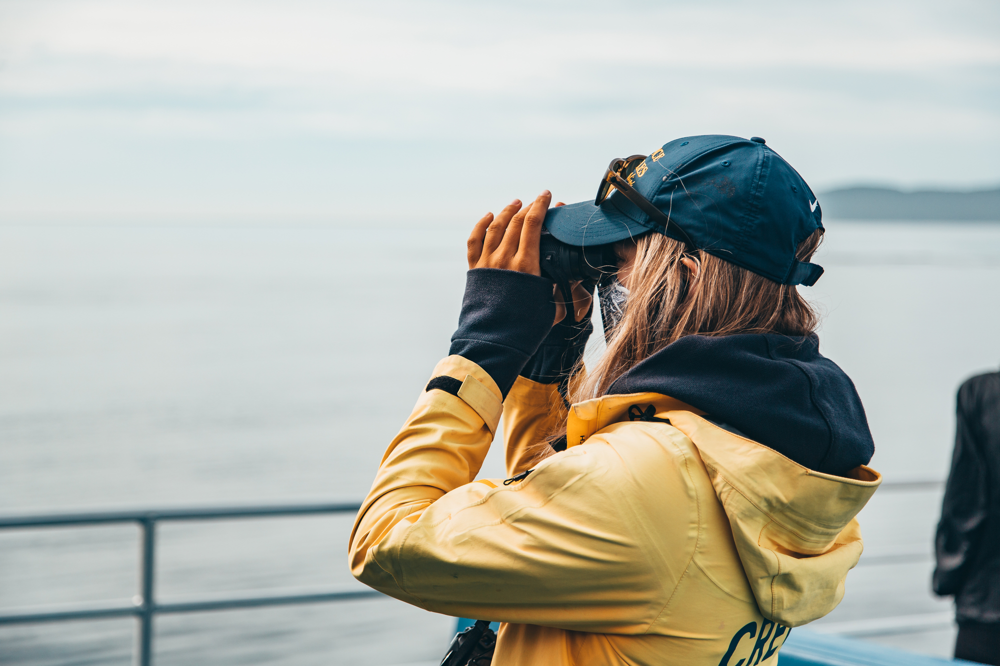 A guide wearing a yellow rain jacket and a blue cap on a Prince of Whales tour scans the water through a pair of binoculars