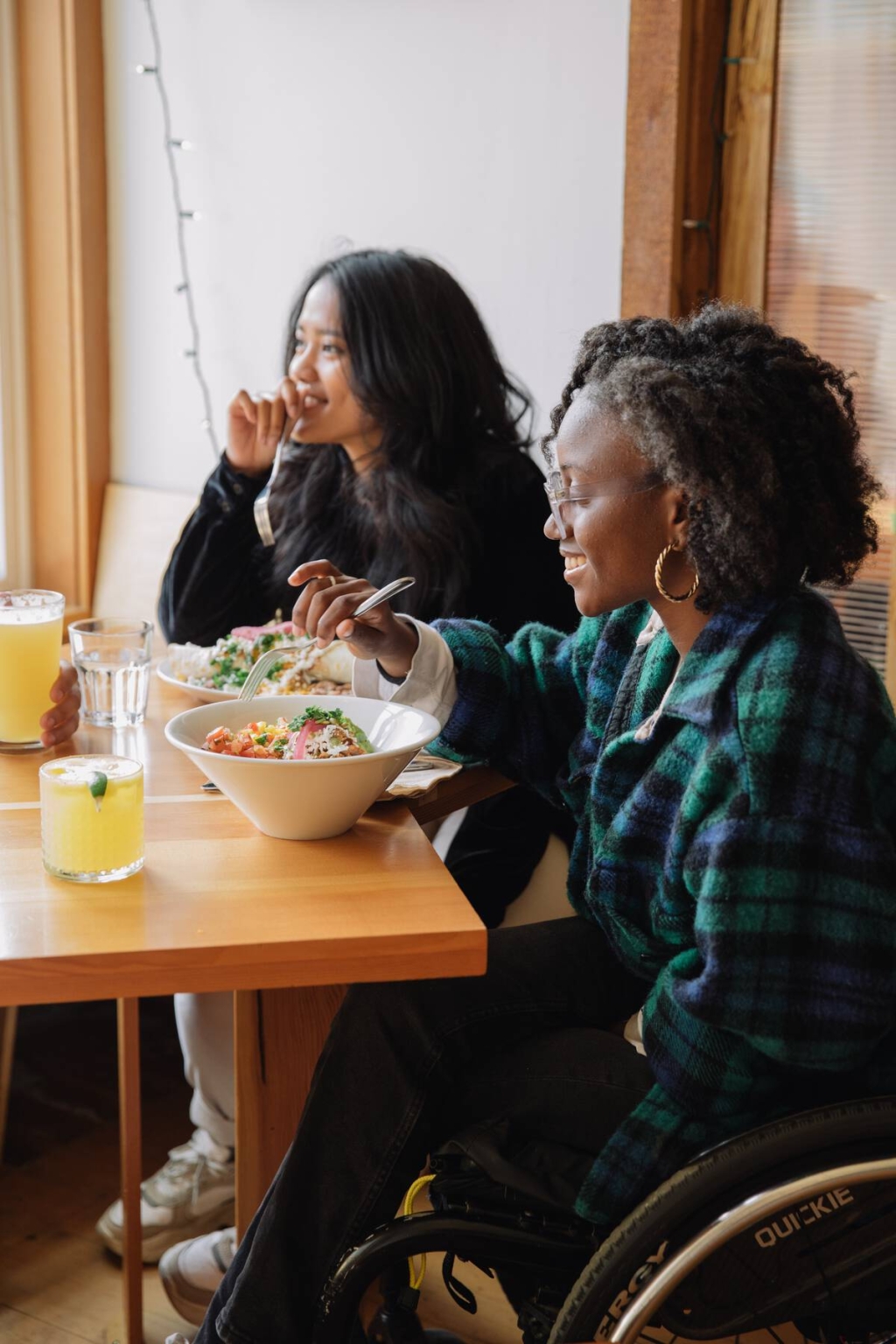 Two people, one in a wheelchair, sit at a wooden table eating bowls of food with drinks on the table.