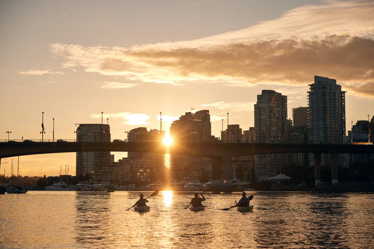Three kayakers paddle toward a bridge at sunset.