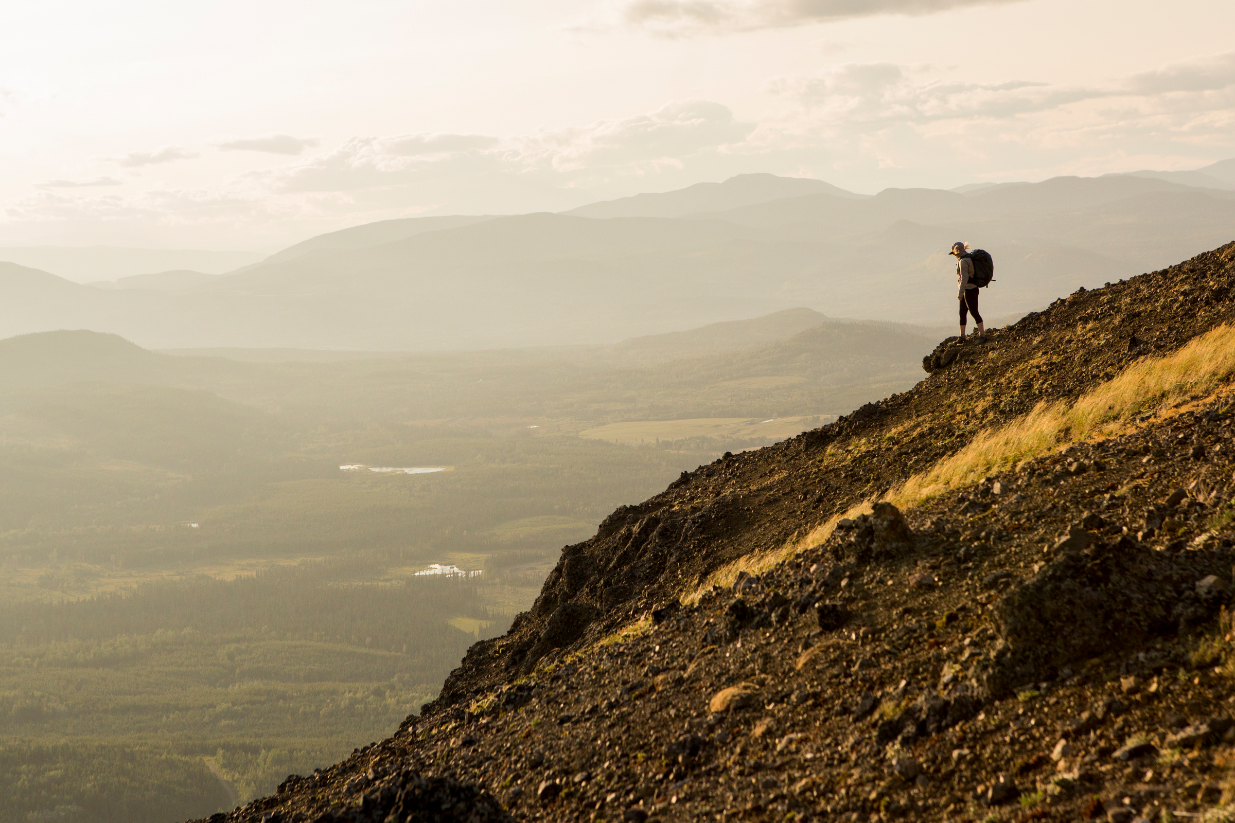 A person with a backpack is perched on a tall mountain overlooking distant mountains and lush valley below.