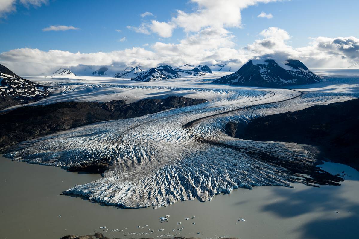 A snaking glacier with white and blue ice snakes across an icefield, dotted by mountains and a glacial lake.