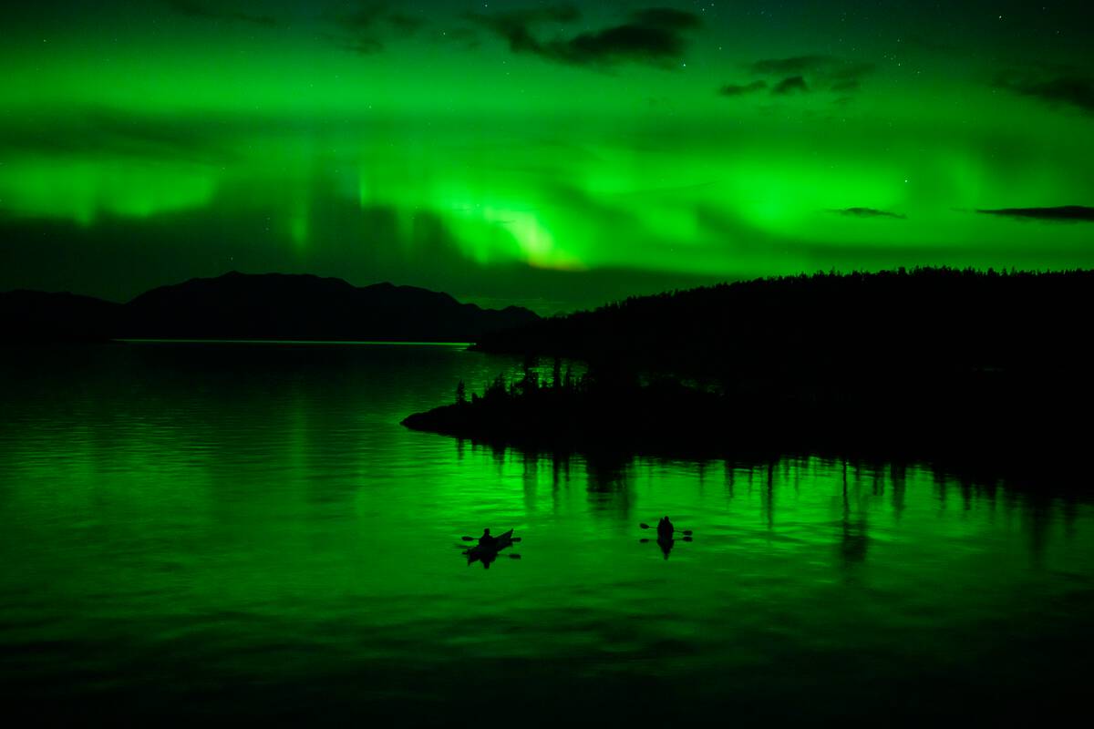 Two people kayak at night under the green glow of the Northern Lights.