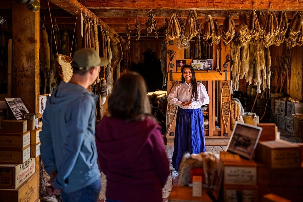 An museum guide stands in a fur trade era building, educating visitors about the heritage and history of the historic site.