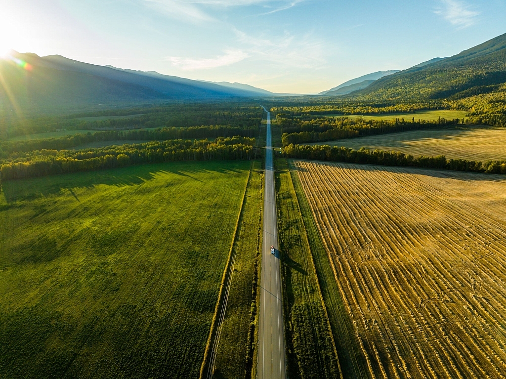 A RV drives along a ribbon of highway that lies between farmland and rugged forest with mountains in the distance.