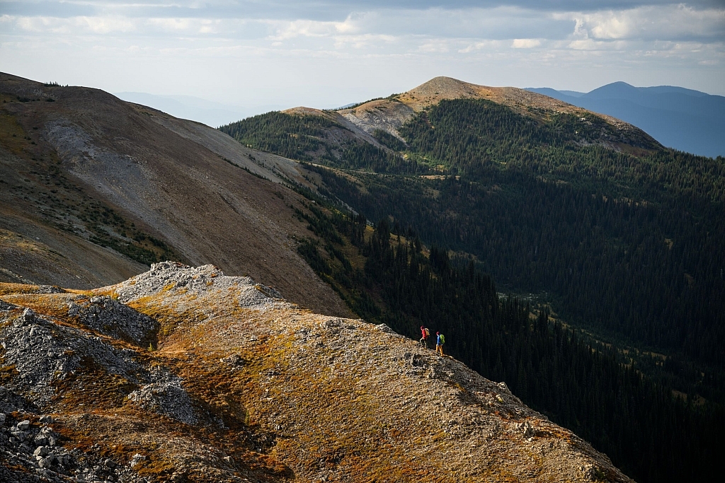 Two people in the distance walk along a rugged ridgeline that overlooks dense forest and orange-tinted mountains.