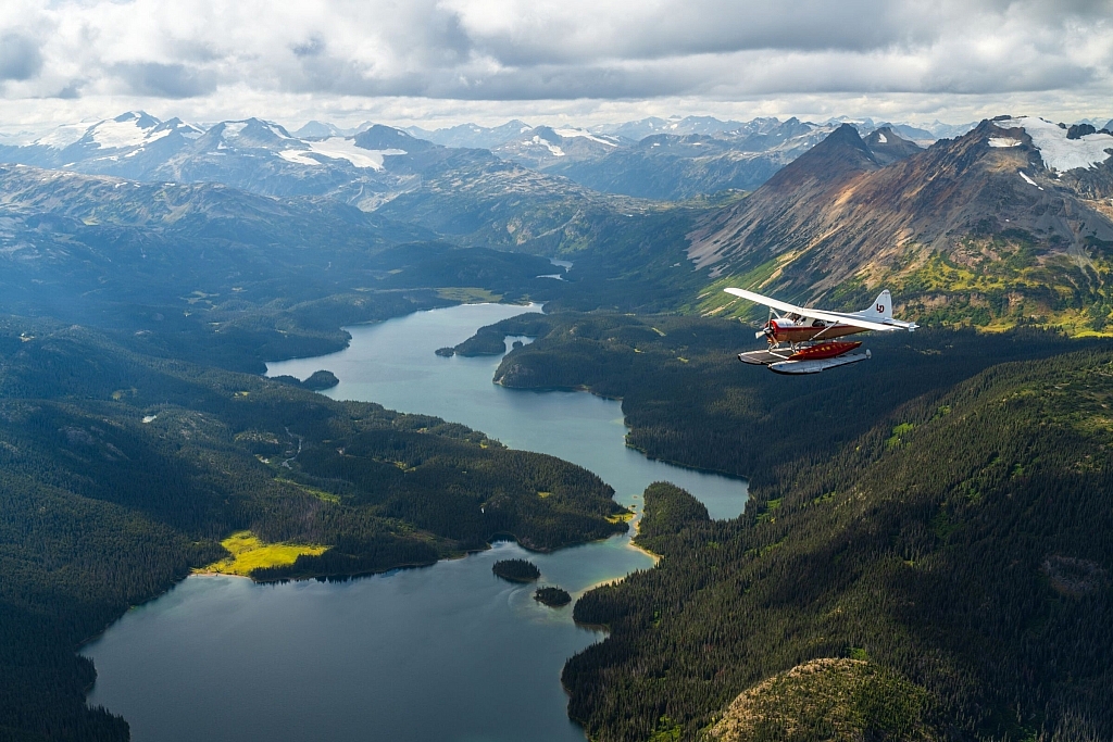 A float plane flies above lakes that are surrounded by dense forest and snow-capped mountains.