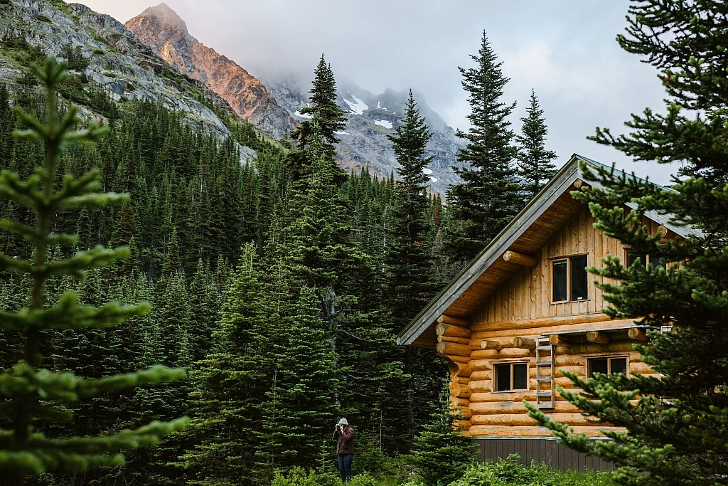 A person stands in front of a log cabin surrounded by rugged mountains and dense forest.