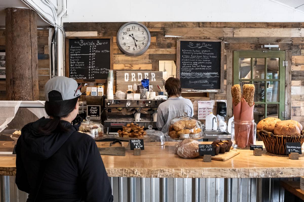 A person waits at the counter of a cafe while a server makes a coffee. The counter is full of baked treats.