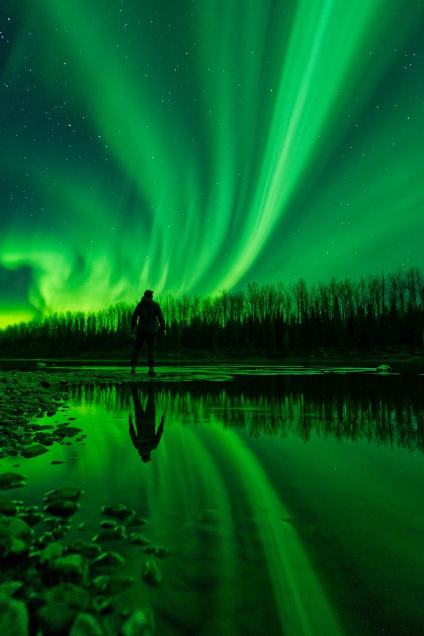 Man stands along the water, looking up at the northern lights.