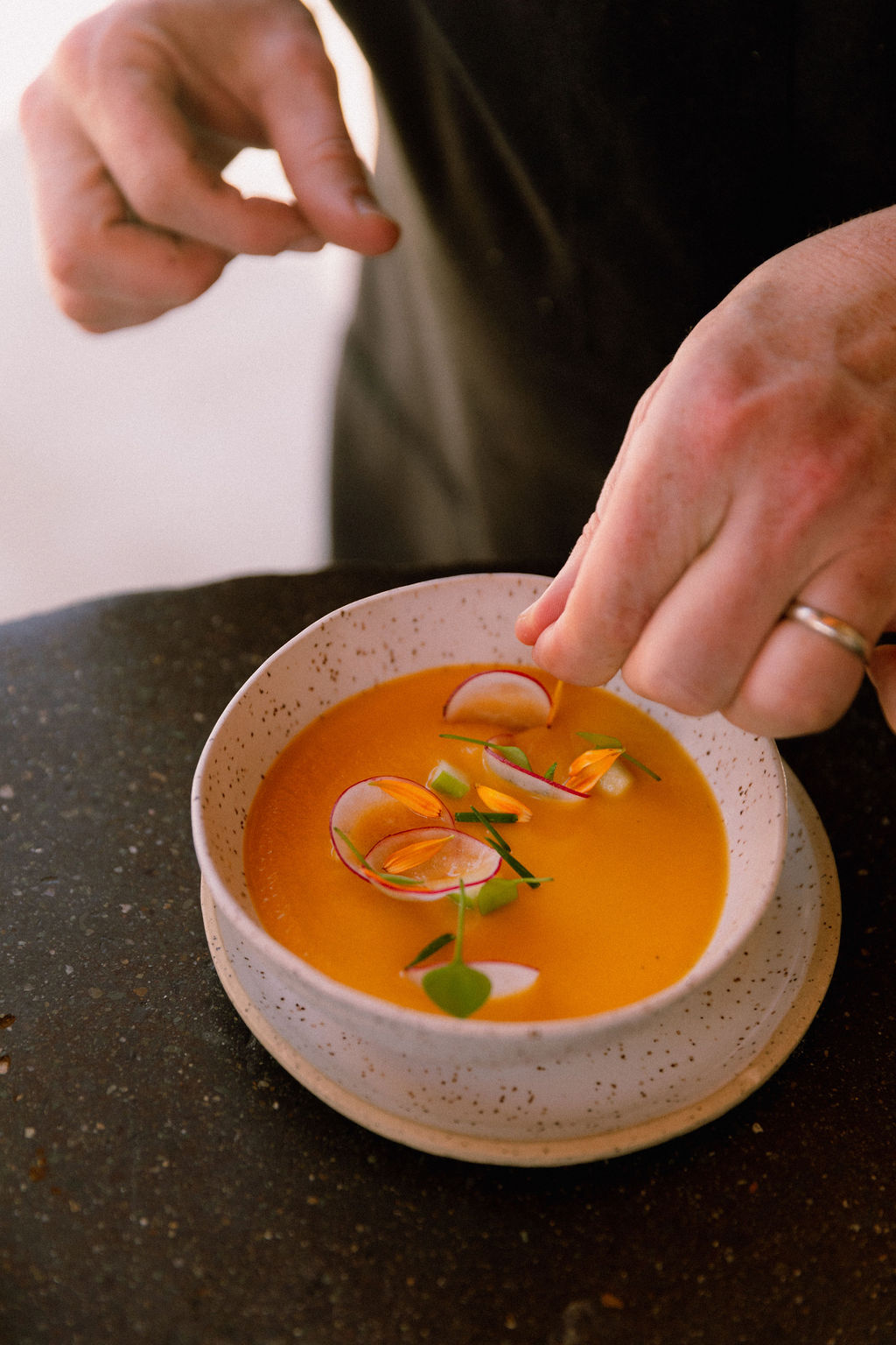 A hand hovers over a bowl of deep orange soup, adding garnishes to the rich dish.