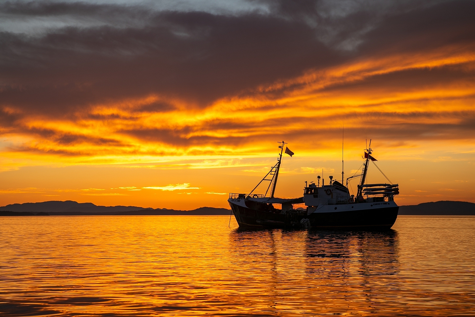 A boat sits on the water, darkened against a mountainous backdrop at sunset