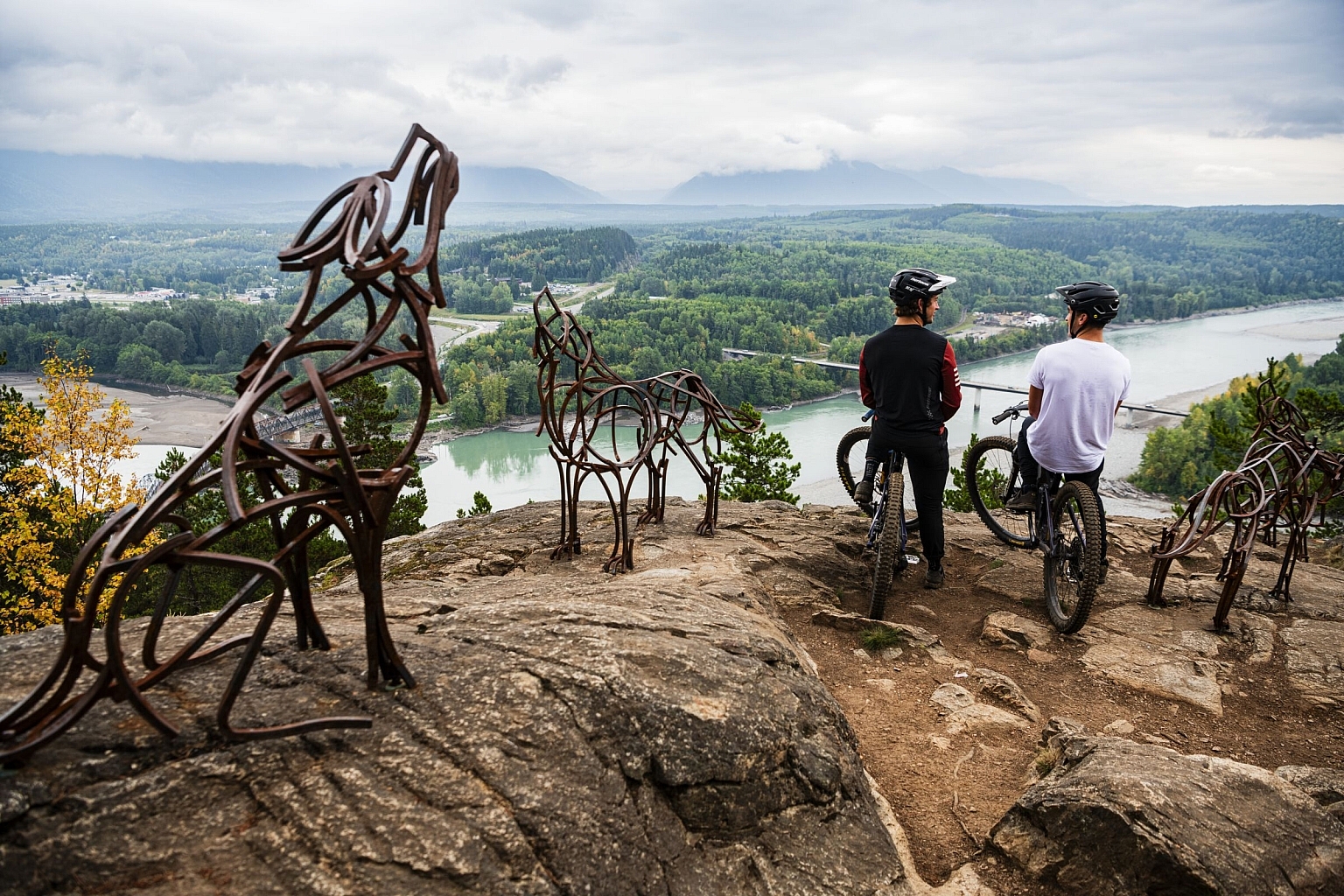 Mountain bikers stand at a lookout surrounded by iron statues of wolves and bears, taking in views of a snaking river and lush forest.