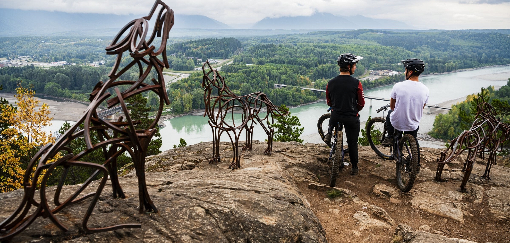 Mountain bikers stand at a lookout surrounded by iron statues of wolves and bears, taking in views of a snaking river and lush forest.