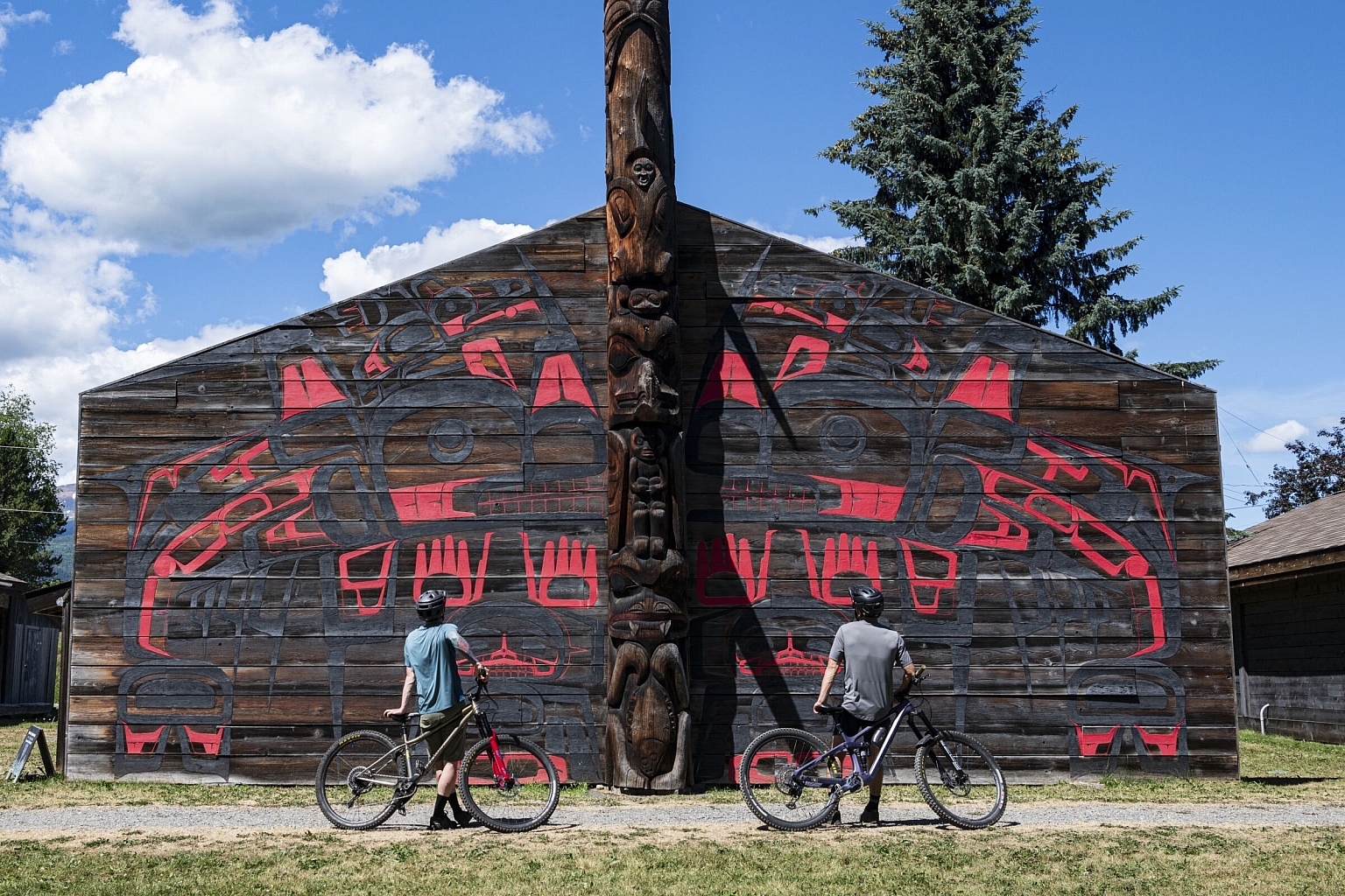 Two mountain bikers wheel their bikes past a longhouse with Indigenous artwork in 'Ksan.