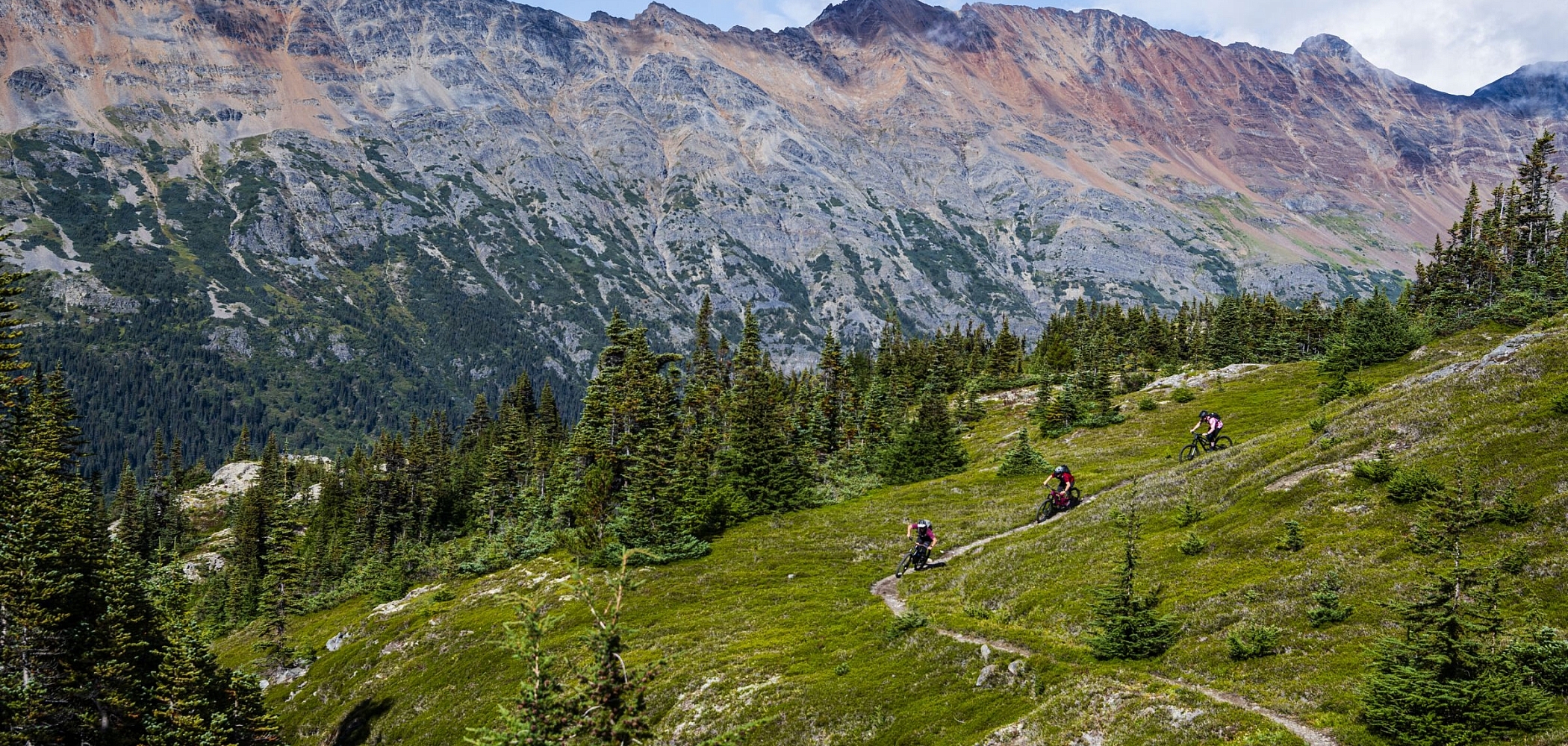 Riders weave their way through a green alpine meadow with jagged peaks in the background.