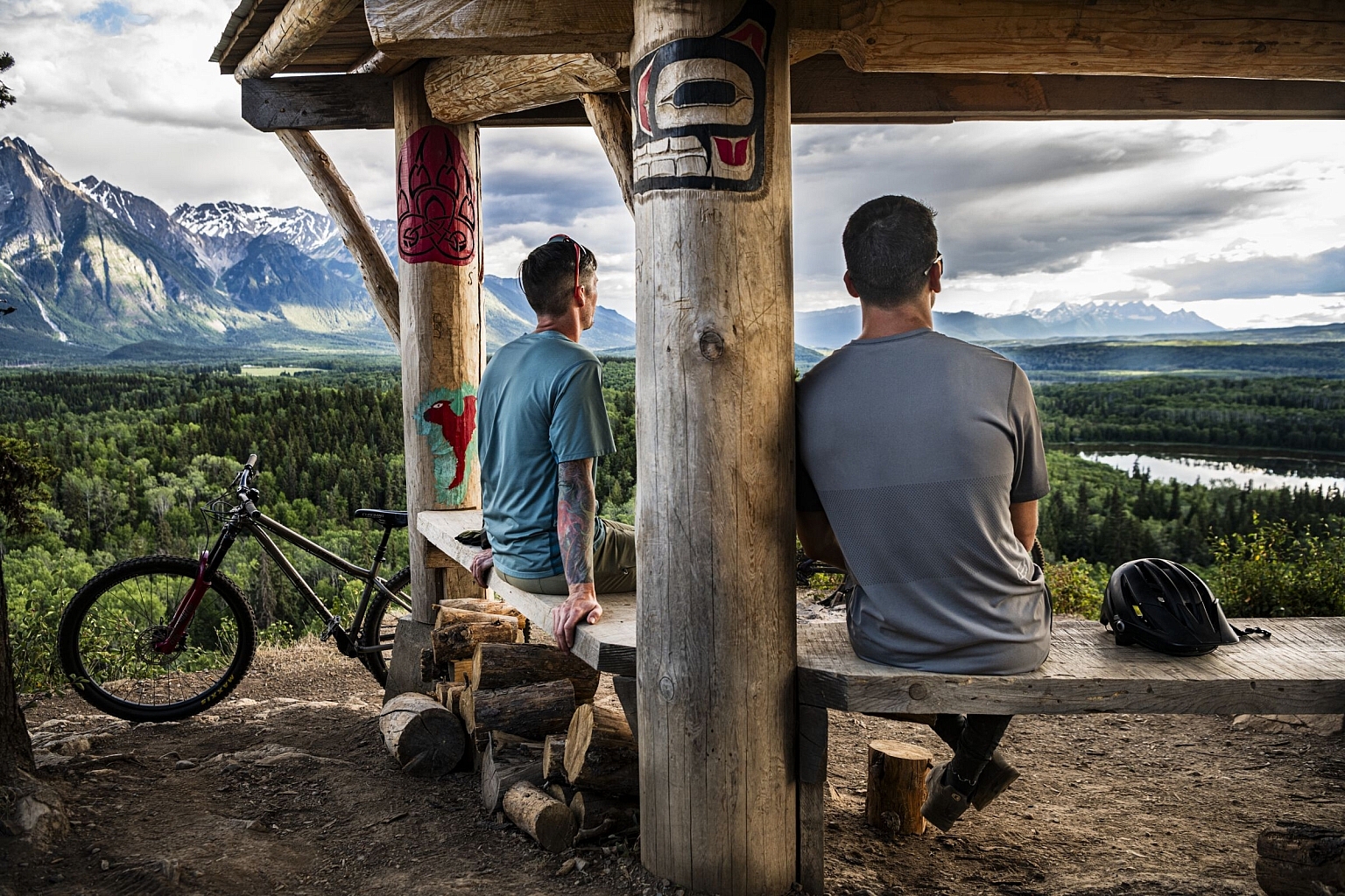 Two mountain bikers sit at an Indigenous-carved wooden bench looking out at a river valley with snow-capped mountains in the background.