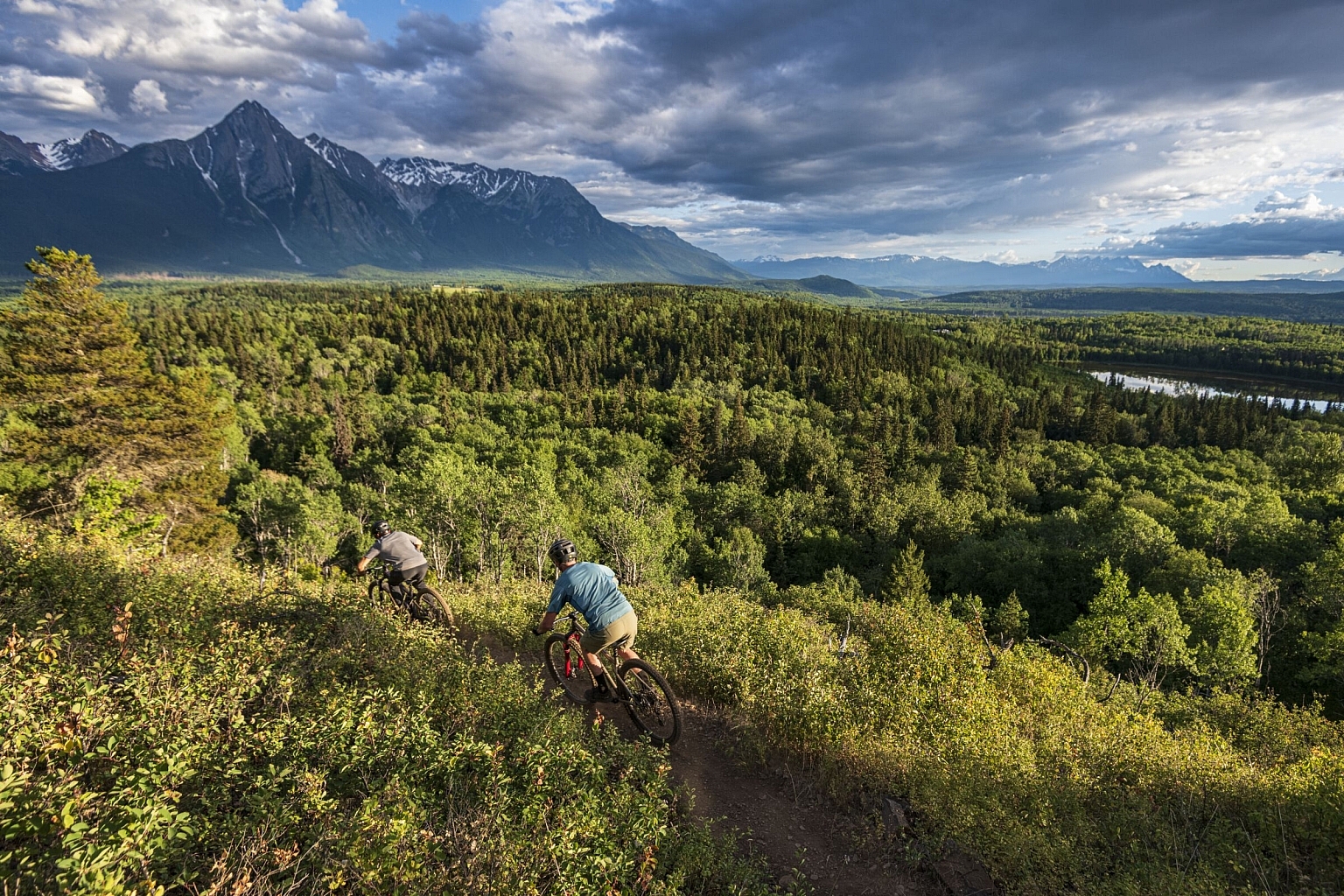 Bikers ride through a verdent trail with a river on the right-hand side and sharp mountains ahead.