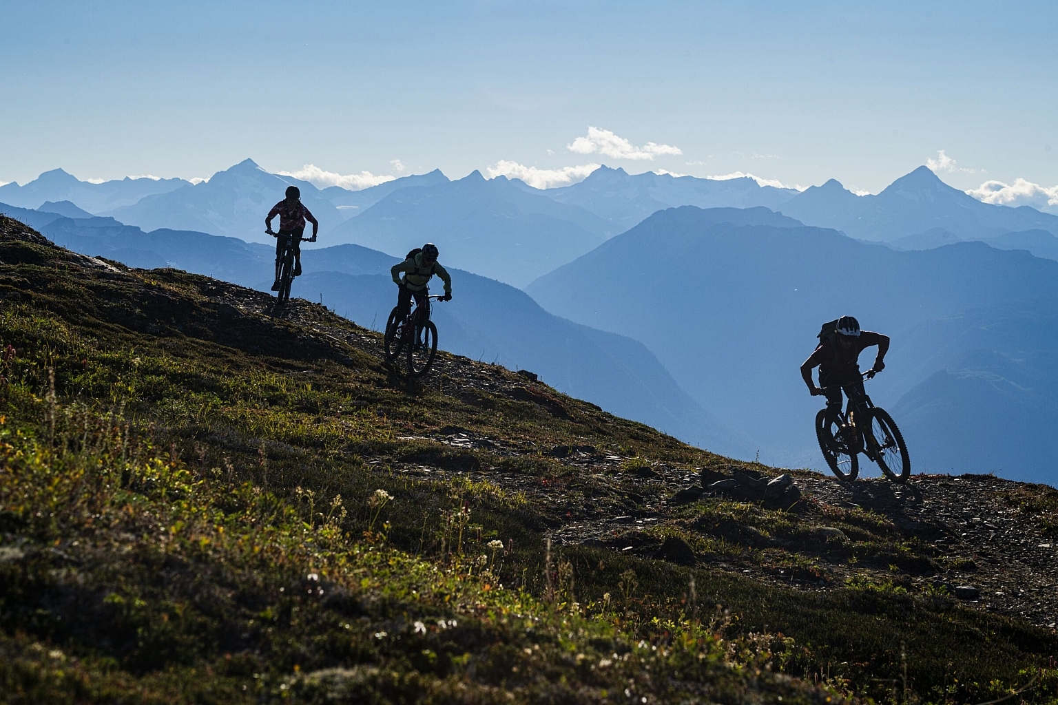 Three mountain bikers descend an alpine trail with green in the foreground and a vast mountain range in the background.