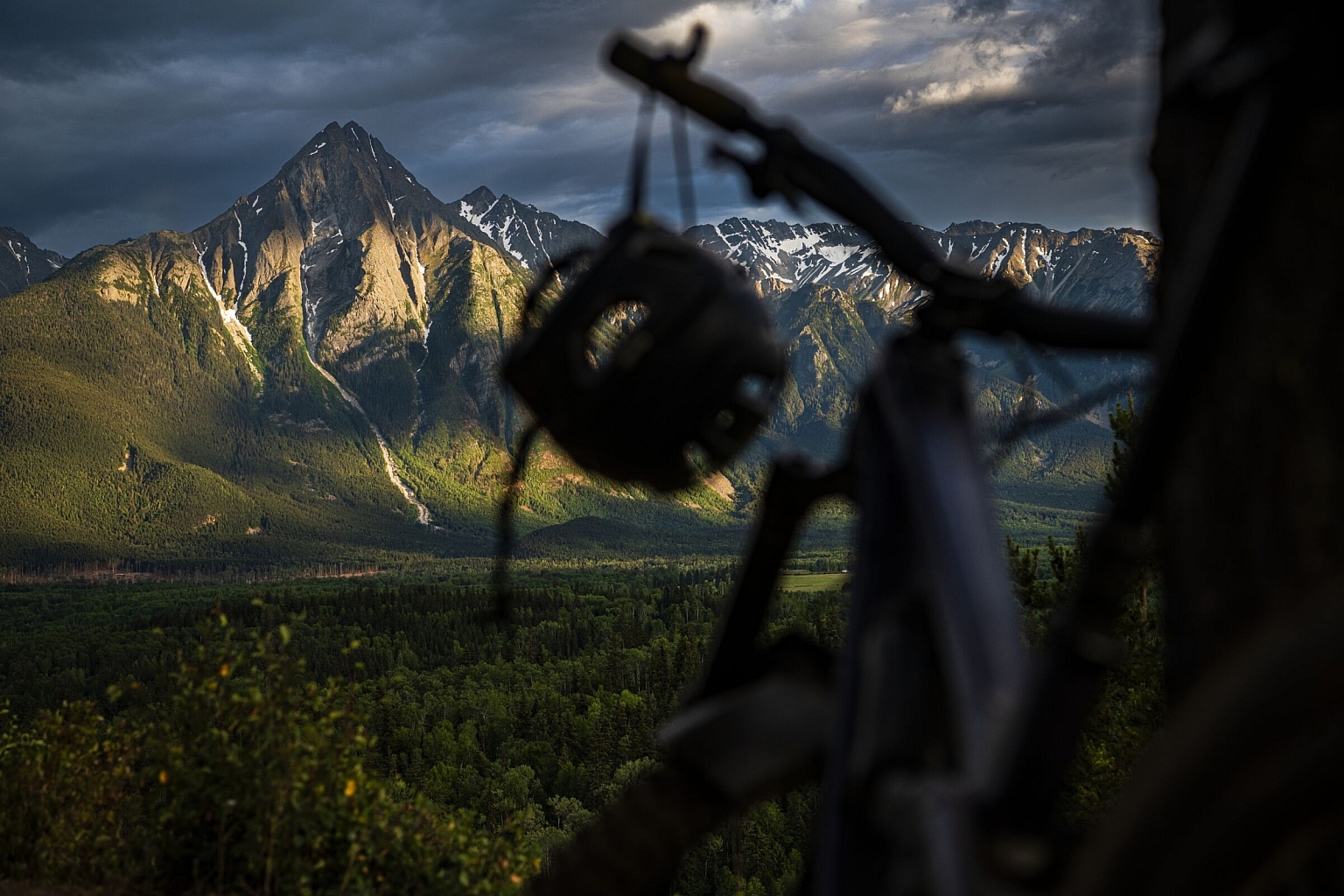 A mountain bike with dangling helmet sits in the foreground with a dramatic mountainscape with lush meadows in the background.