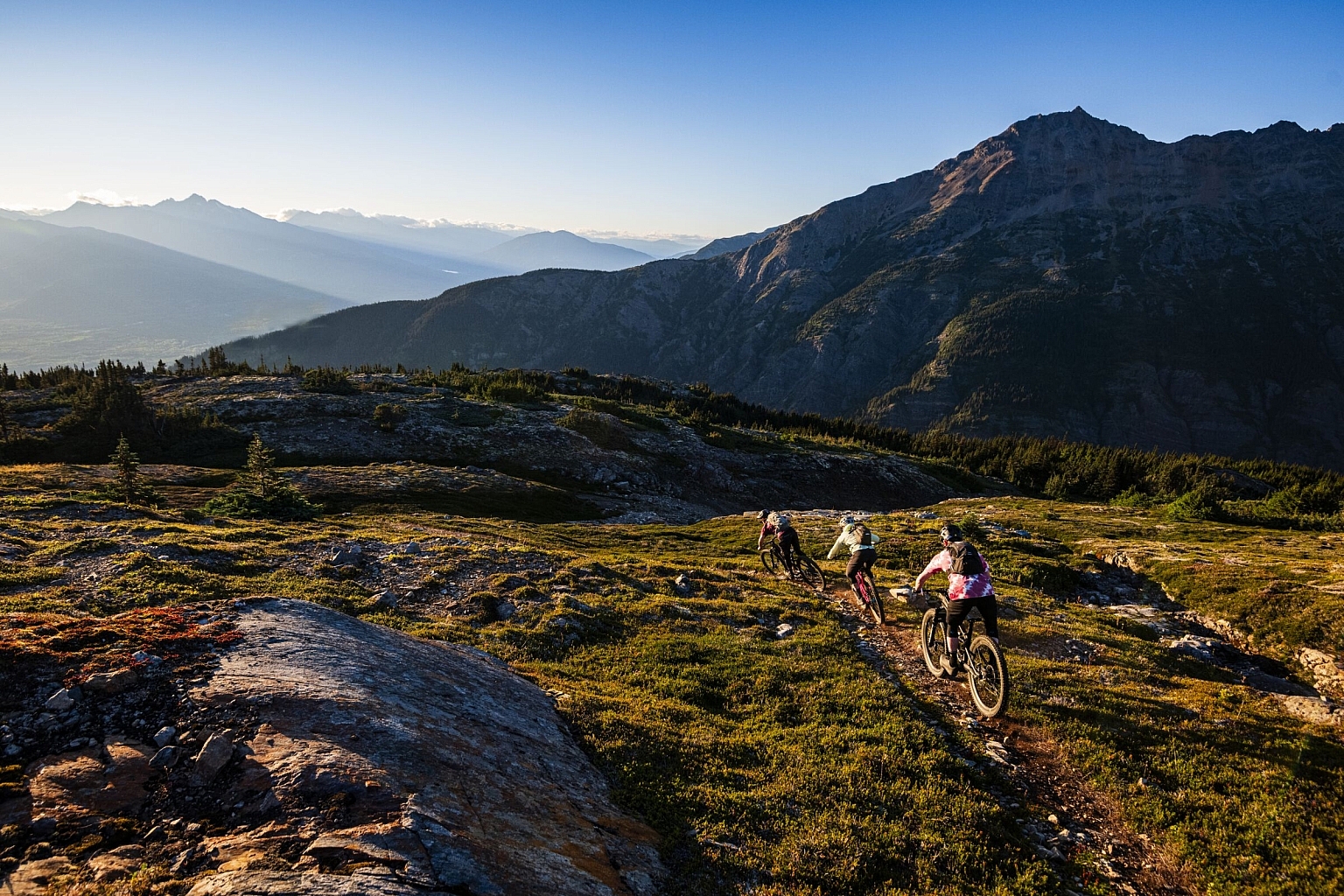 A group of mountain bikers rides through an alpine trail with green voliage and craggy mountains.