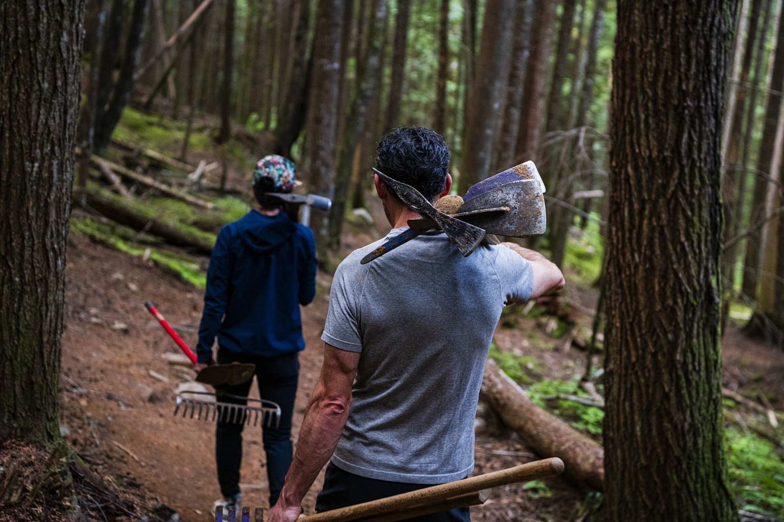 Two trail building volunteers walk along a trail with tools over their shoulders.