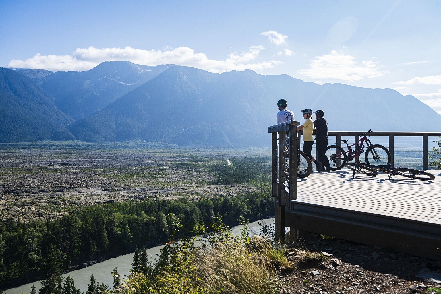 Two people on a wooden viewing platform, with bikes parked to the side, look out over a vast river valley.