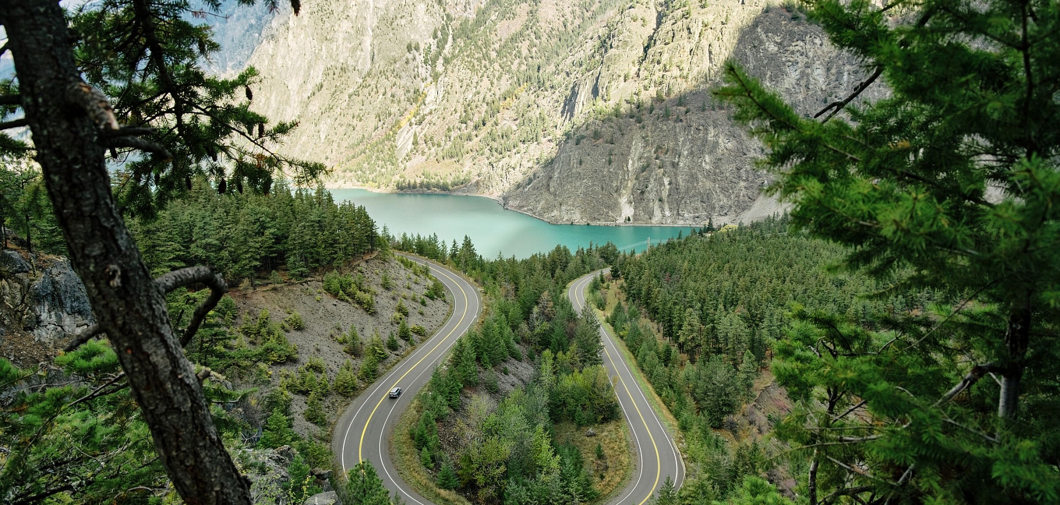 Vehicle travelling along a snaking highway with a turquoise lake and towering mountains in the background.